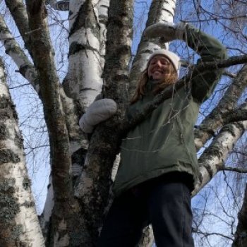 Woman wearing mittens and hat stood in a birch tree looking out